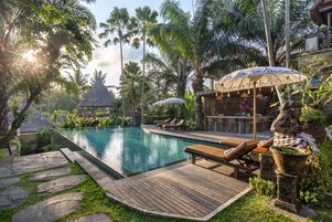 Un área de piscina con sillas de descanso y sombrillas, rodeada de vegetación tropical y una zona de bar.