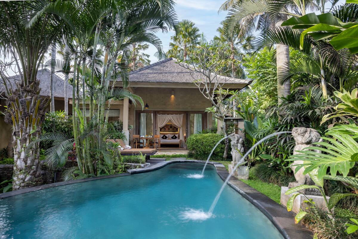 A pool with a waterfall, surrounded by lush tropical plants and a building with a covered seating area.