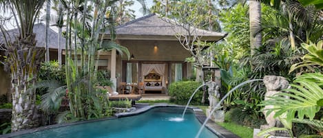 A pool with a waterfall, surrounded by lush tropical plants and a building with a covered seating area.