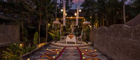 A tropical outdoor dining area with a table set for two, surrounded by palm trees and a decorative pathway with candles.
