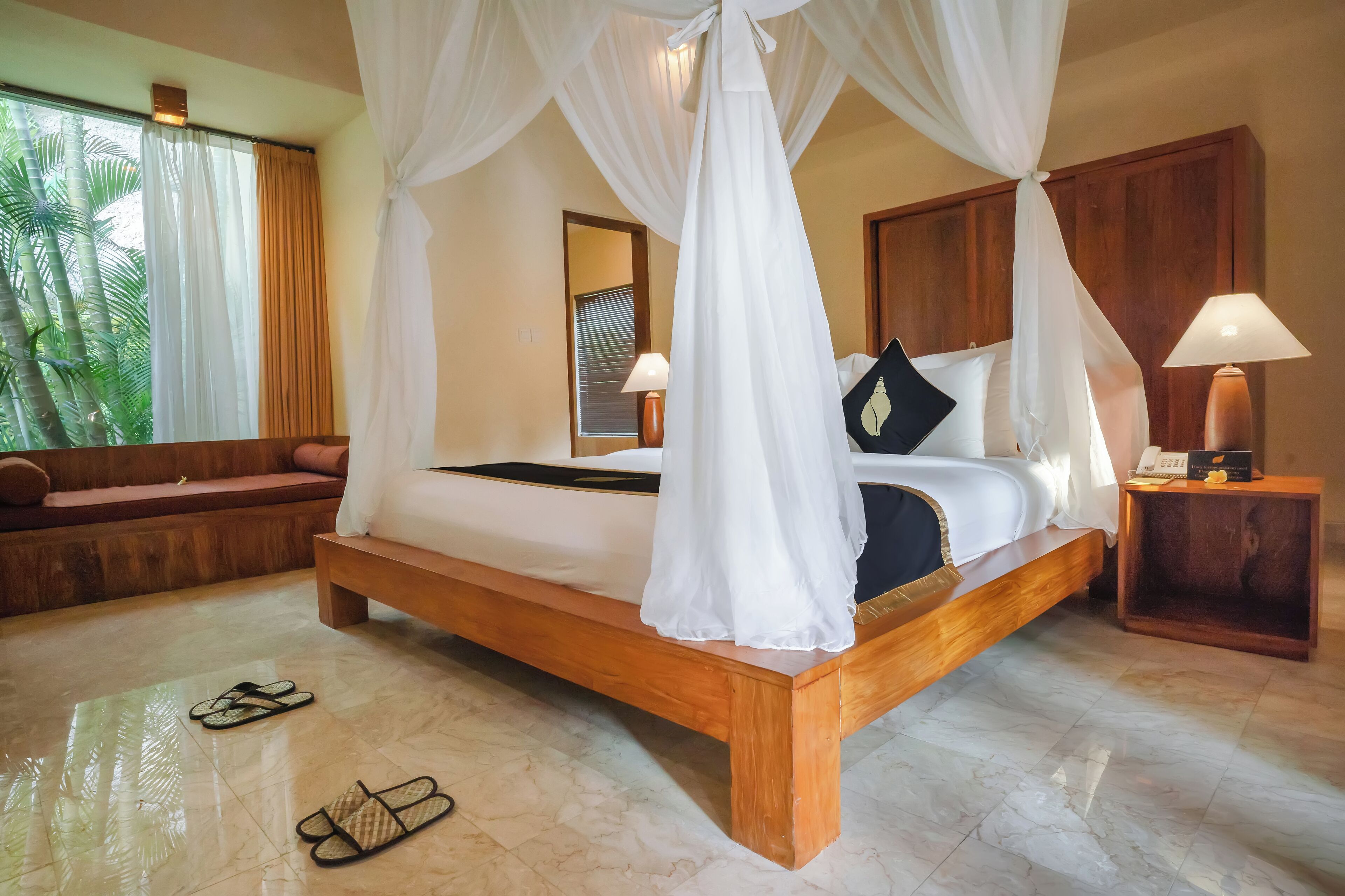 Un dormitorio amplio con una cama de madera, dosel blanco y vistas a palmeras a través de amplias ventanas.