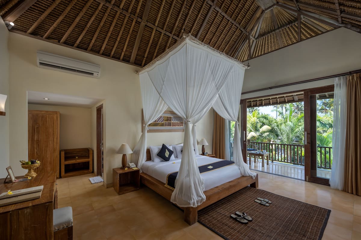 A spacious bedroom with a large bed, a wooden desk, a chair, and a balcony with a view of palm trees.