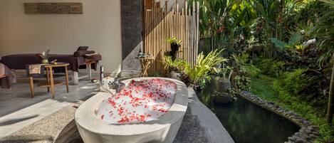 A spa area with a stone bathtub filled with water and rose petals, a massage table, and a small table with a plant.