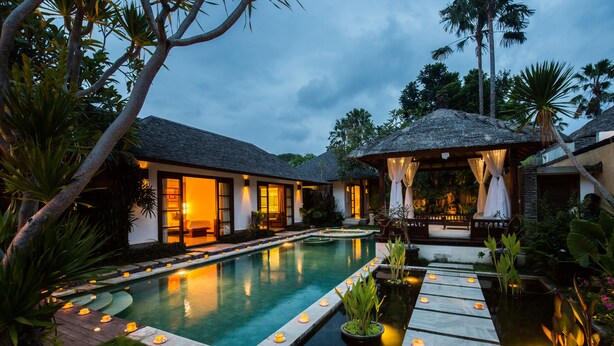 A pool with a wooden deck, surrounded by greenery and a house with large windows. - Villa Seriska Satu Sanur Bali (Denpasar)