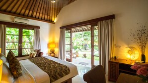A bedroom with a bed, bedside tables, a desk, and a view of a garden through large windows. - Villa Seriska Satu Sanur Bali (Denpasar)