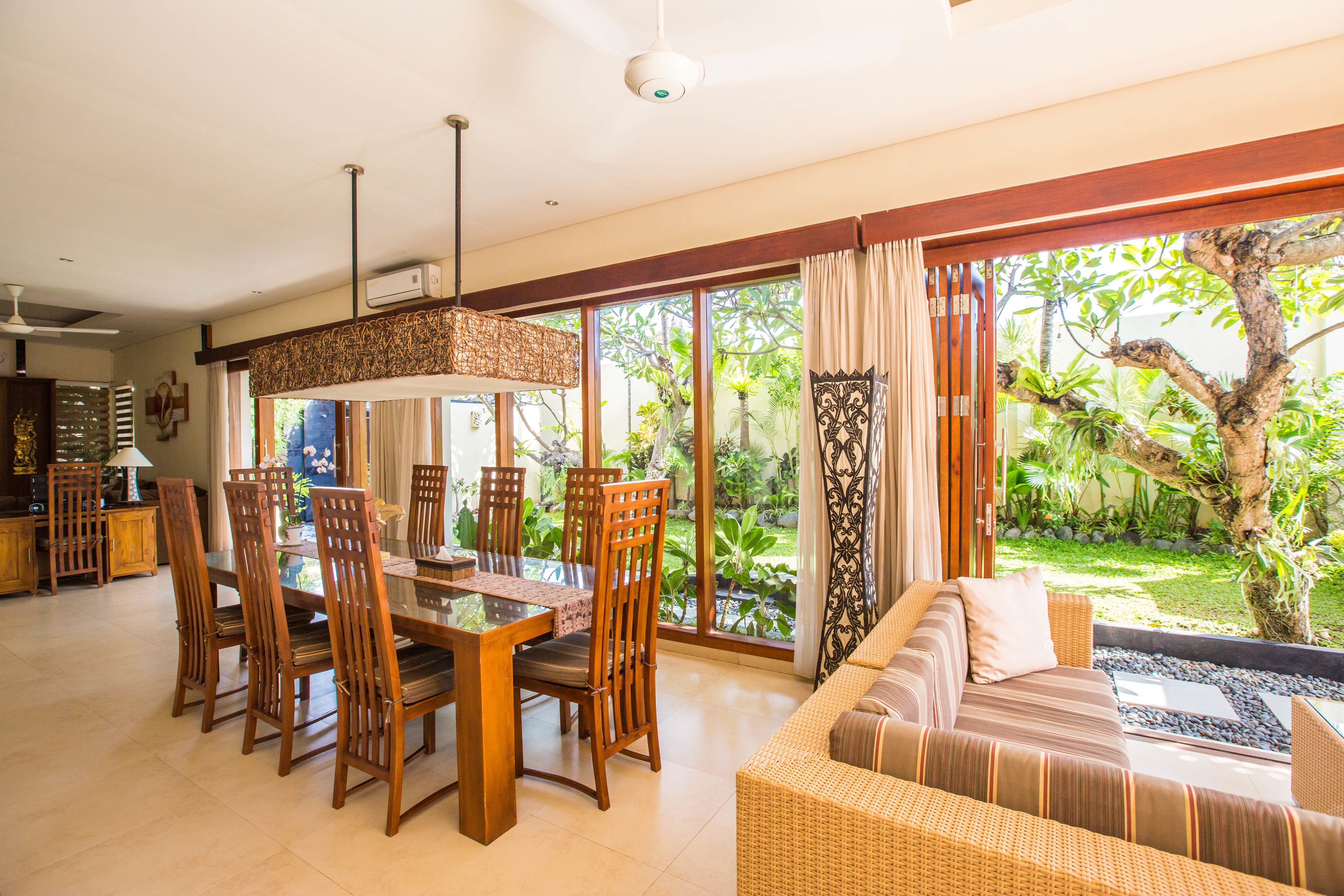 Uma sala de estar ampla com uma mesa de jantar e cadeiras, um sofá e janelas grandes que oferecem vista para um jardim exuberante.