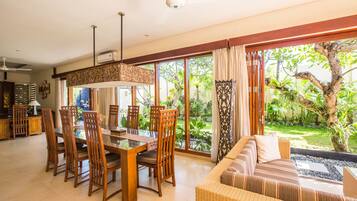 Uma sala de estar ampla com uma mesa de jantar e cadeiras, um sofá e janelas grandes que oferecem vista para um jardim exuberante.