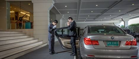 Un hombre con traje abre la puerta de un coche en un aparcamiento cubierto, con una escalera y la entrada de un edificio al fondo.