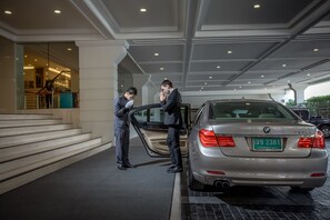 A man in a suit opening a car door in a covered parking area with a staircase and a building entrance in the background.