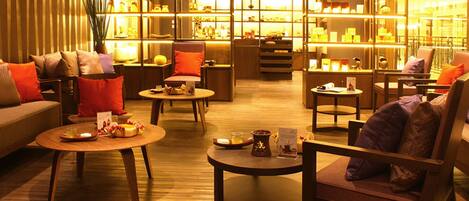 A warmly lit lounge area with sofas, armchairs, and tables, featuring a variety of decorative items and a bookshelf in the background.