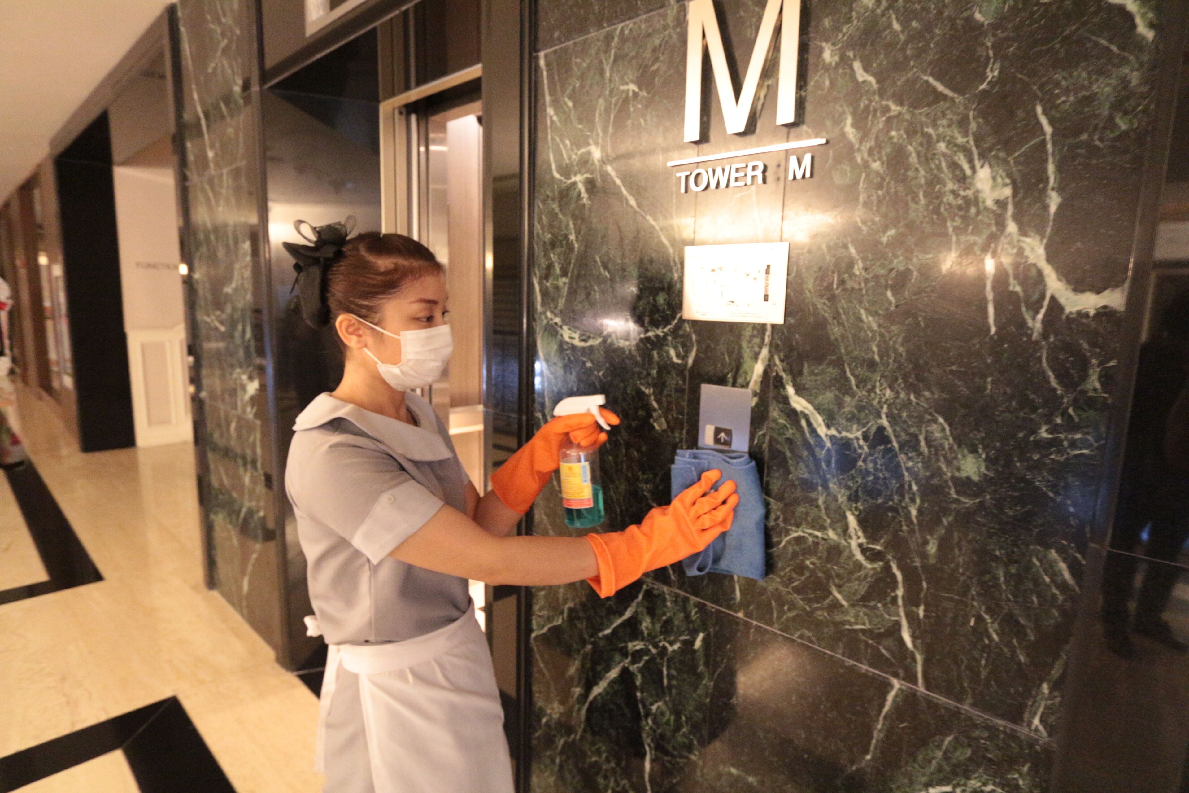 A person cleaning a marble wall with a spray bottle and cloth in a hallway with a black and white floor pattern.