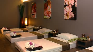 Ein Spa-Raum mit drei Betten, jedes mit Handtüchern und einer Blume. An den Wänden hängen Blumenbilder und eine hohe, grüne Pflanze.