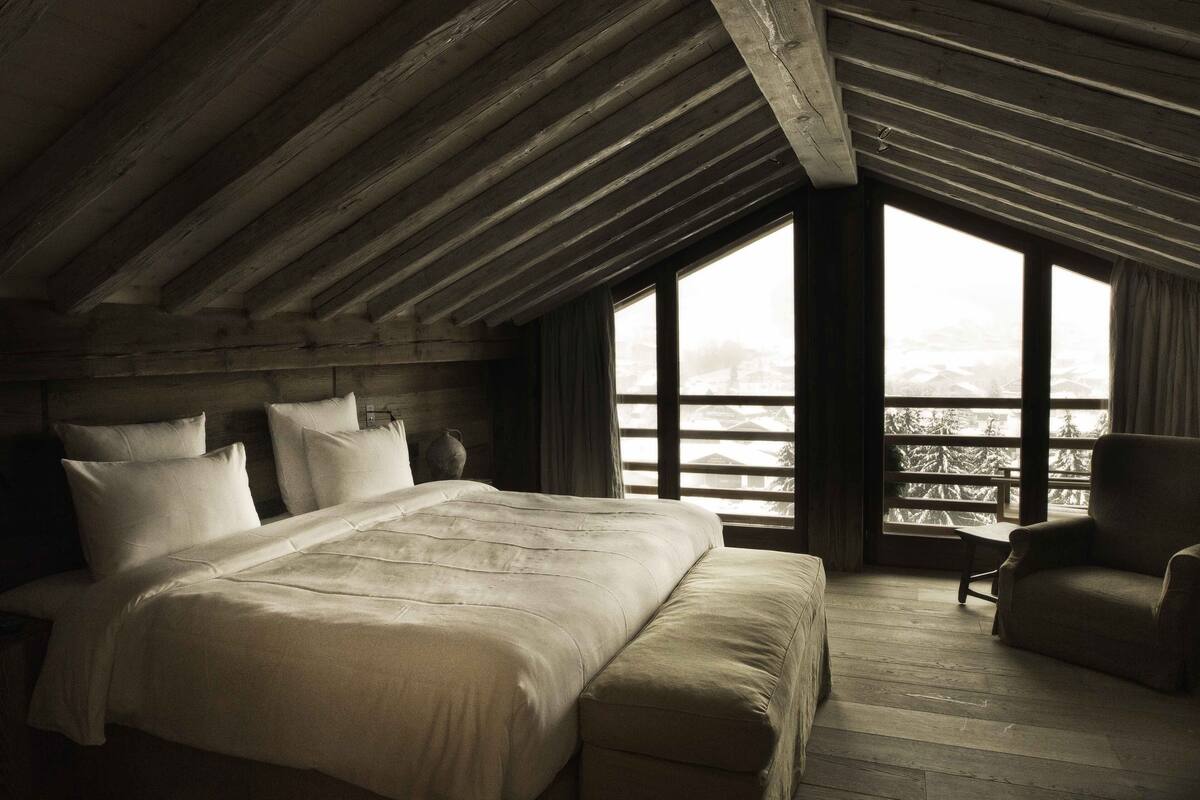 Un dormitorio con una cama grande, una silla y vistas de árboles cubiertos de nieve a través de una ventana.