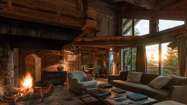 A cozy log cabin interior with a fireplace, wooden furniture, and large windows offering a view of the outdoors. - Zannier Le Chalet (Megeve)