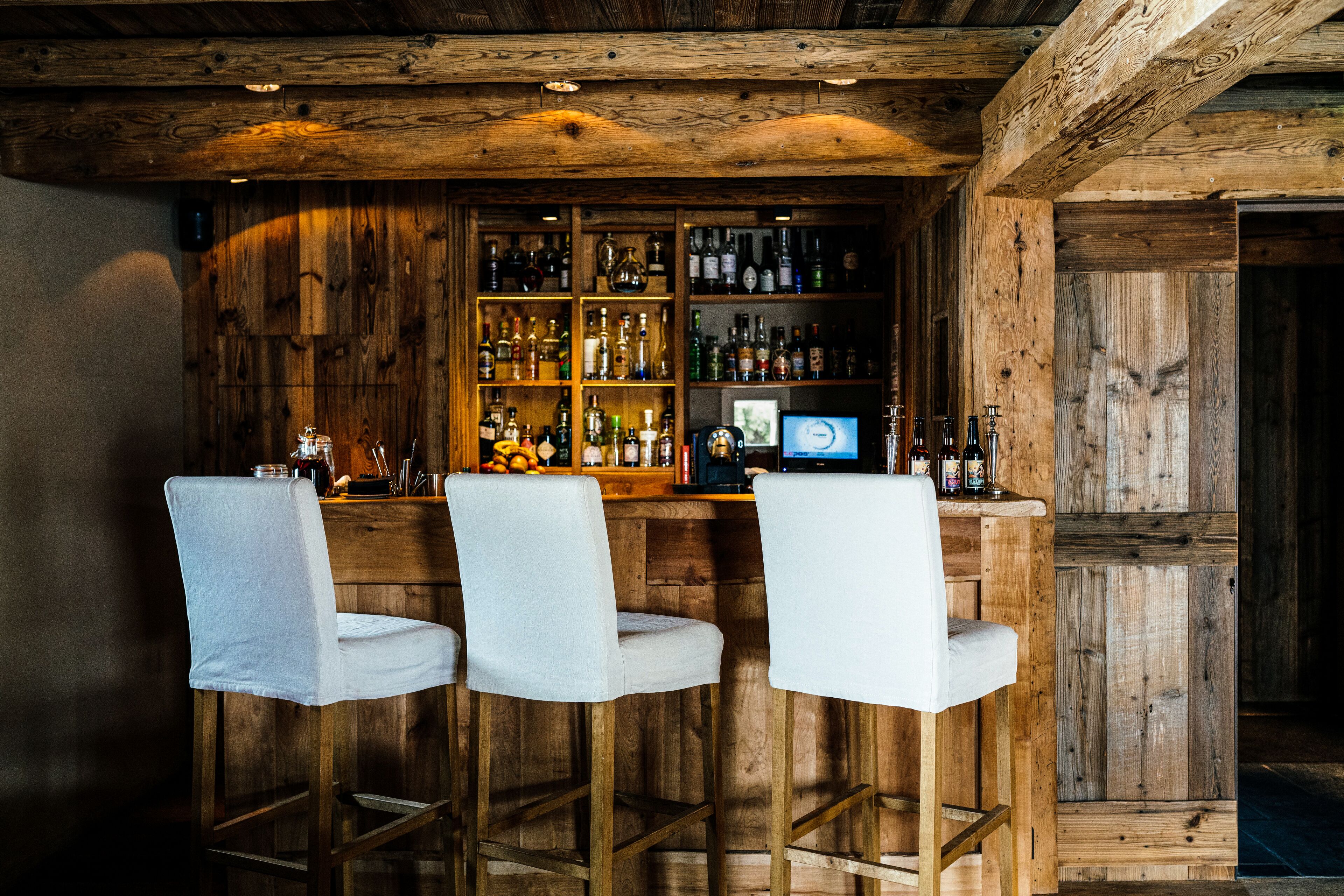 Un coin bar au style rustique, avec des murs en bois et trois tabourets de bar blancs.
