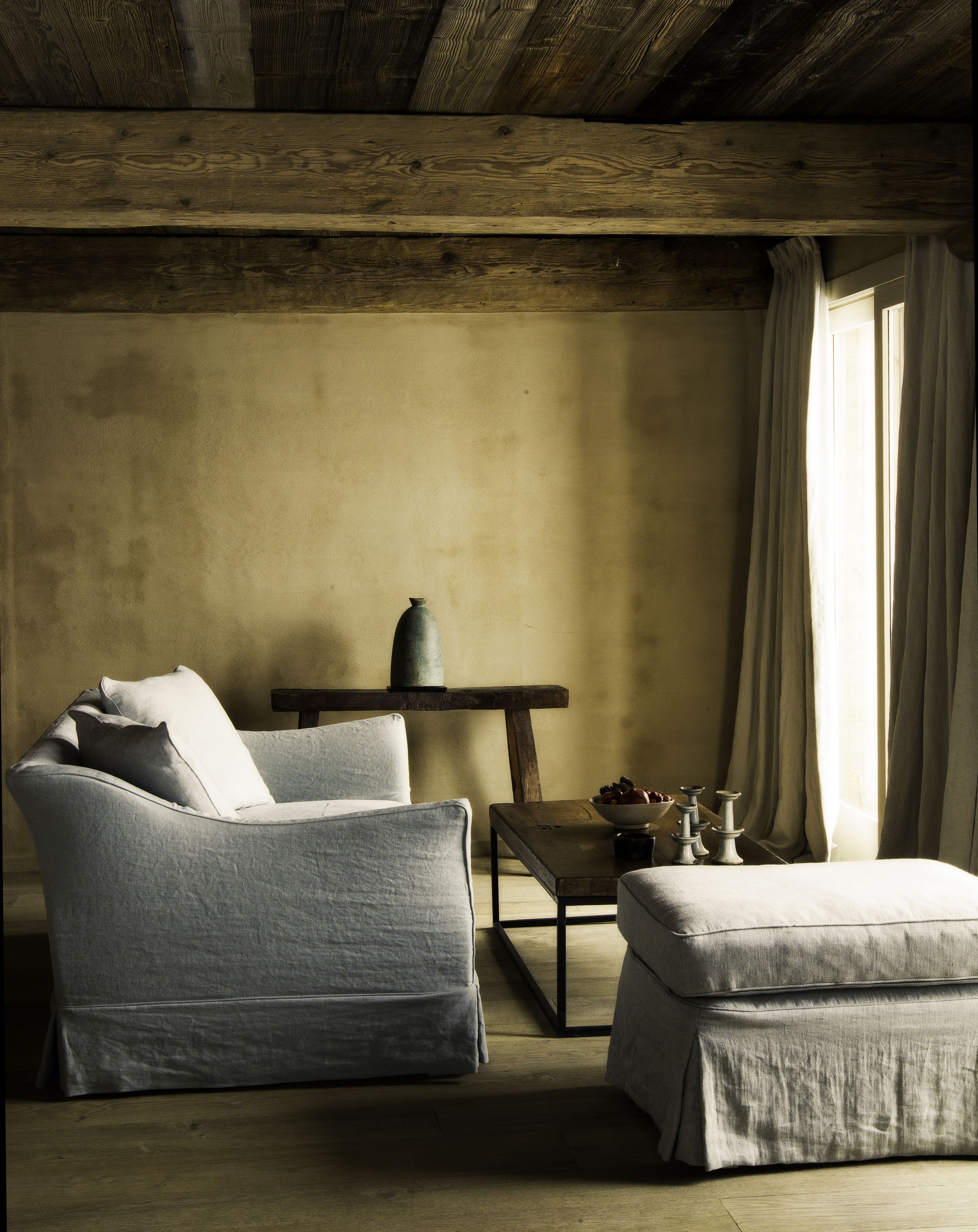 A room with a wooden ceiling, a grey armchair with cushions, a small table with a vase and candles, and a window with curtains.