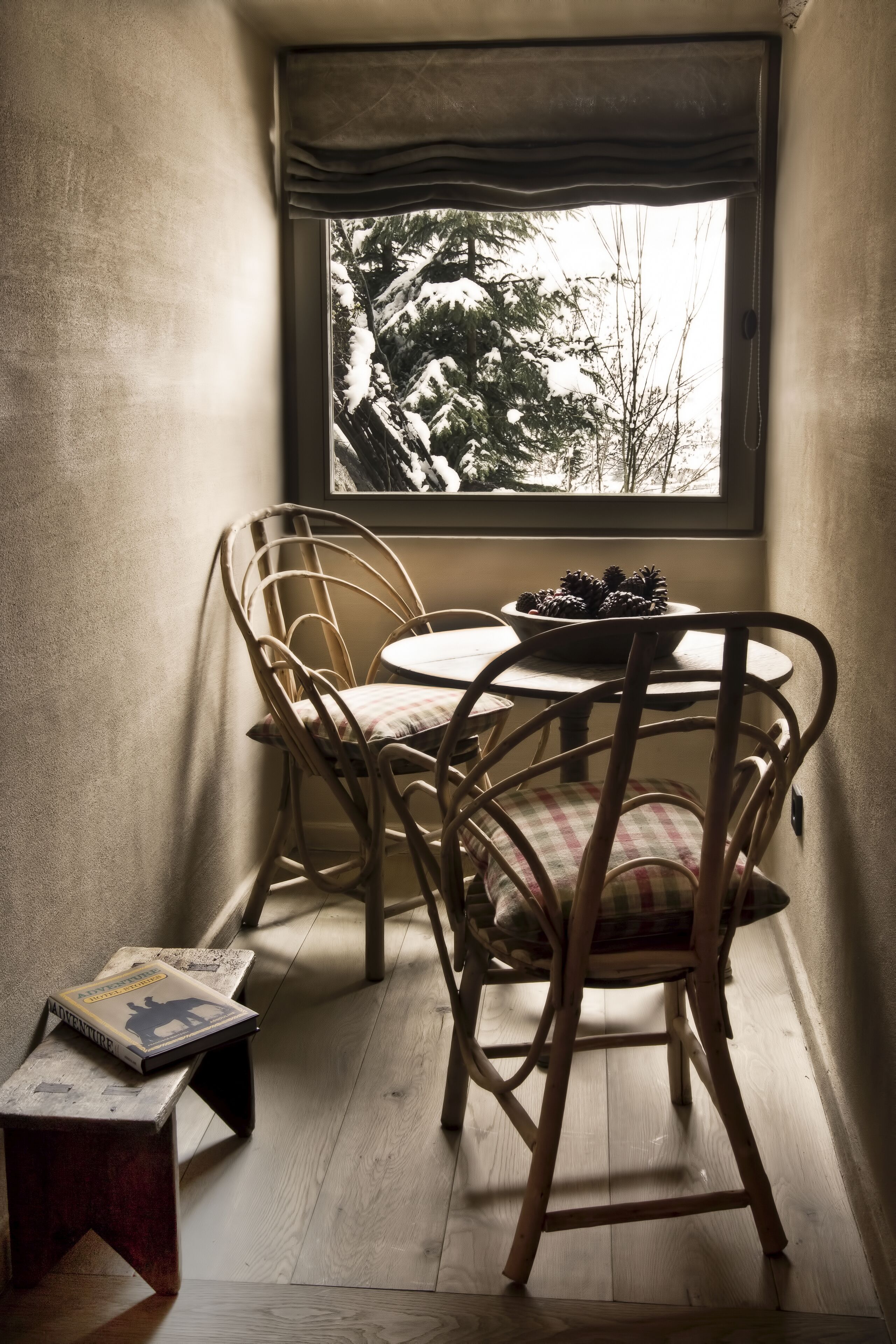 Ein Eckzimmer mit Fensterblick auf eine verschneite Landschaft, einem runden Tisch mit Stühlen und einem kleinen hölzernen Hocker mit einem Buch.