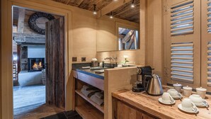 A rustic bathroom with a wooden vanity, a sink, and a mirror. There is a fireplace in the background, and towels are neatly folded on a shelf. - Zannier Le Chalet (Megeve)