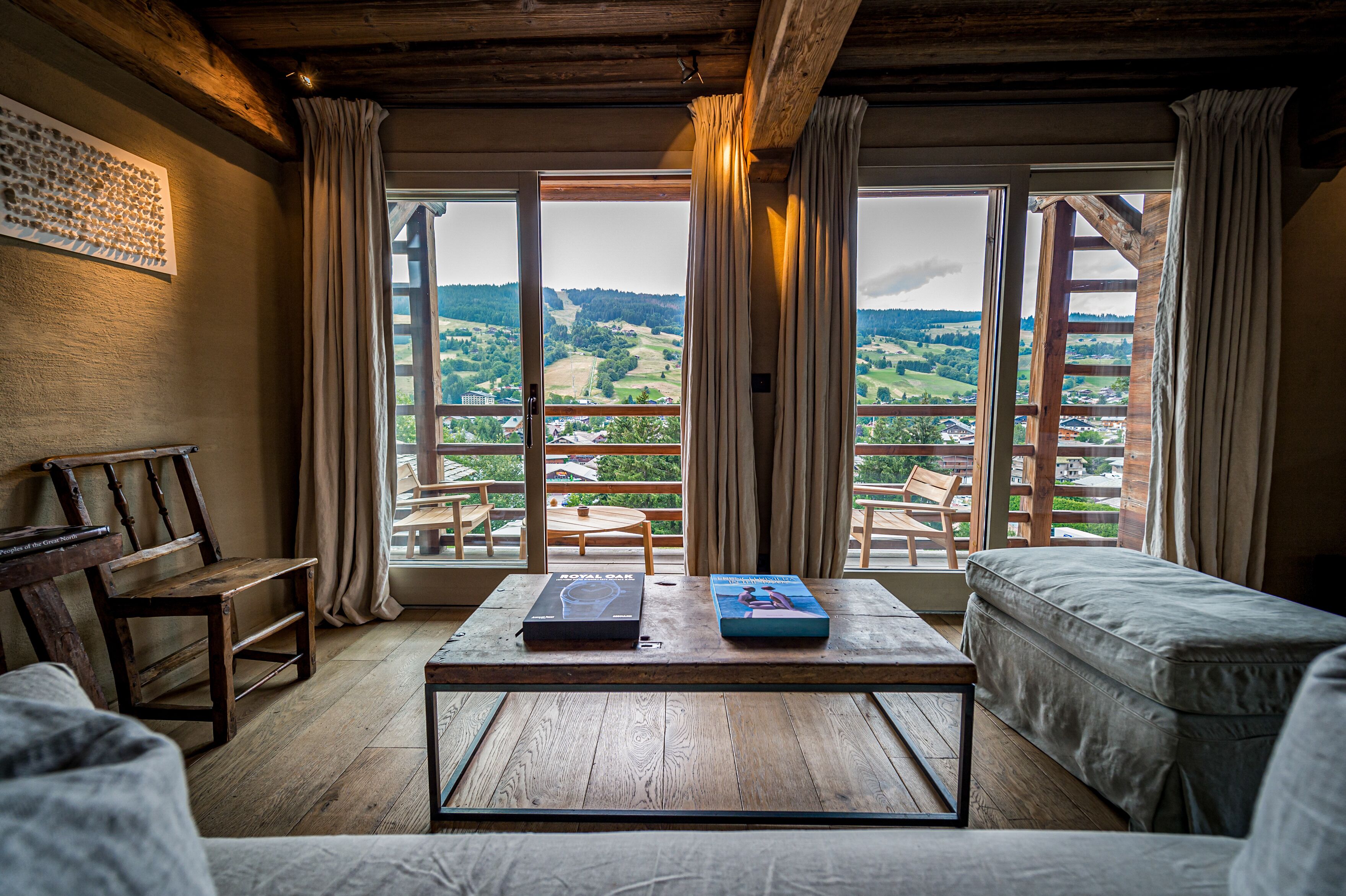 A room with a wooden floor, a table with books, a sofa, and a view of a green landscape through large windows.