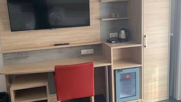 Una habitación moderna con televisión de pantalla plana, un escritorio de madera, una silla roja, un refrigerador pequeño y un armario.
