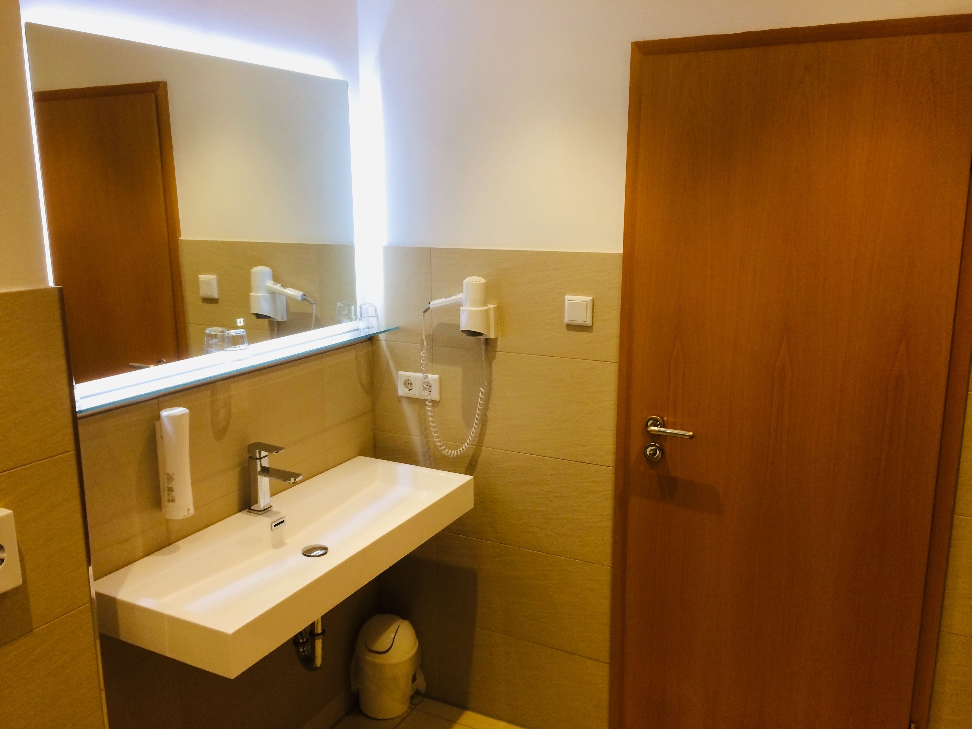 Un baño con lavabo, espejo, secador de pelo y cubo de basura.