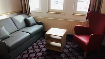 Una habitación con un sofá gris, un sillón rojo y una pequeña mesa de madera. Hay tres ventanas con persianas, y se ve un radiador en la pared.
