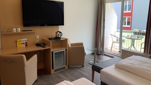 A hotel room with a bed, a desk, a television, a mini-fridge, and a balcony with a table and chairs. - Adesso Hotel Göttingen (Goettingen)