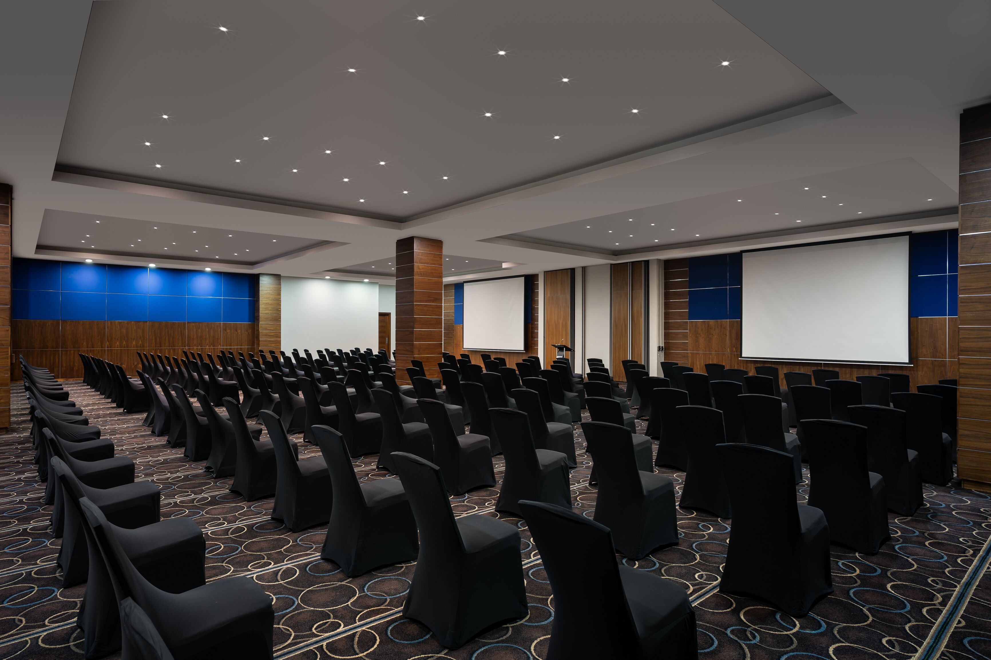 Un salón de conferencias con filas de sillas negras, una pantalla grande y una pared decorativa azul.