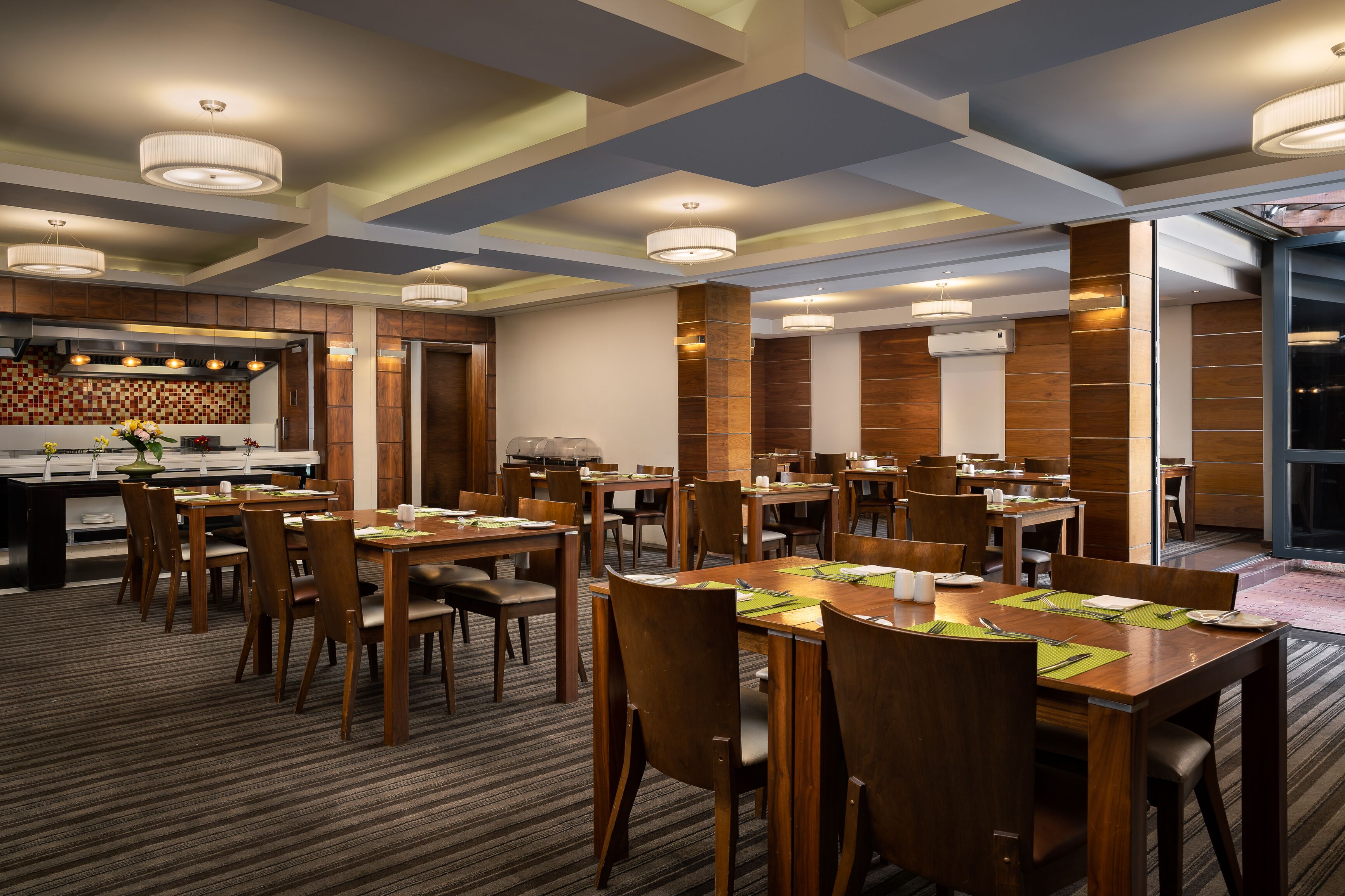 A hotel dining area with multiple tables and chairs, a kitchen in the background, and a modern ceiling design.