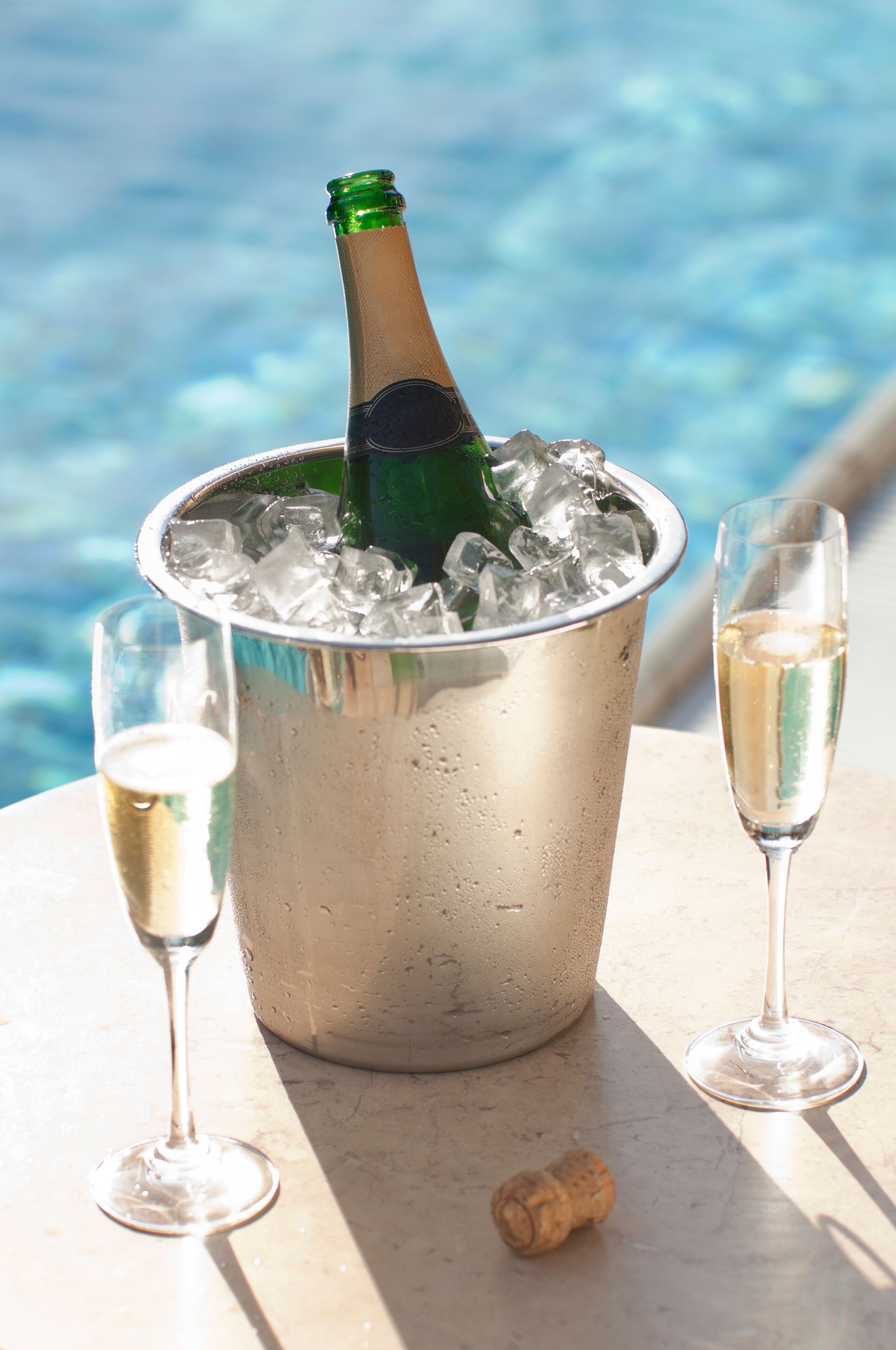 A metal bucket filled with ice and a champagne bottle, two glasses of champagne, and a cork on a table by a pool.