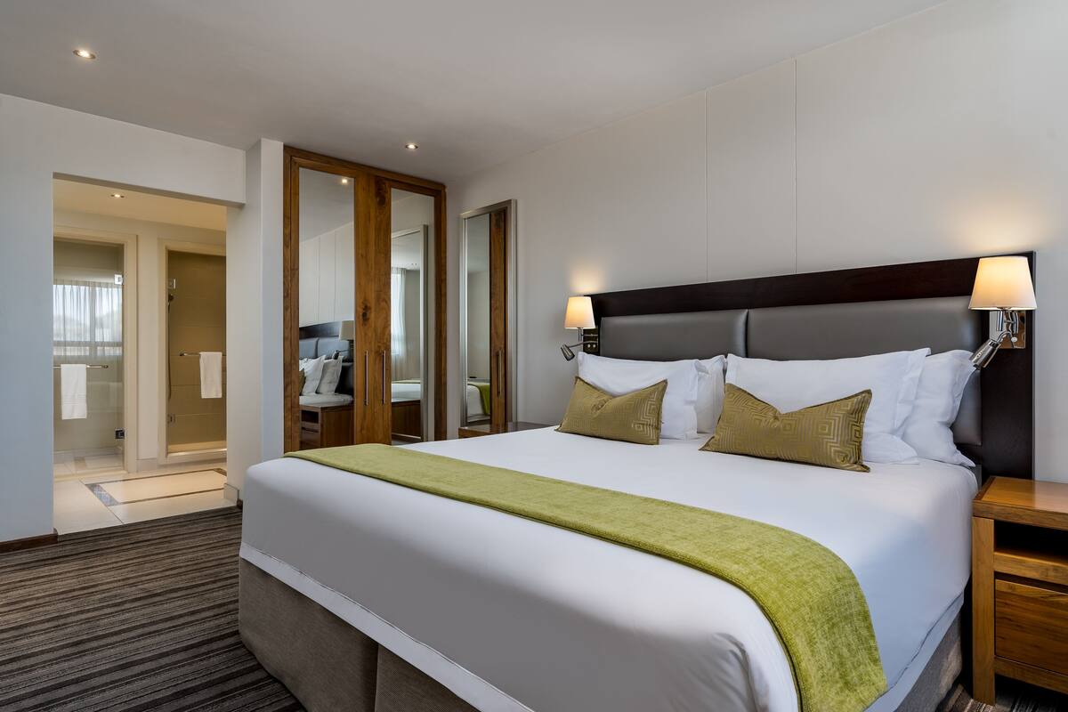 A hotel room with a large bed, a wooden headboard, two bedside lamps, and a green throw blanket.