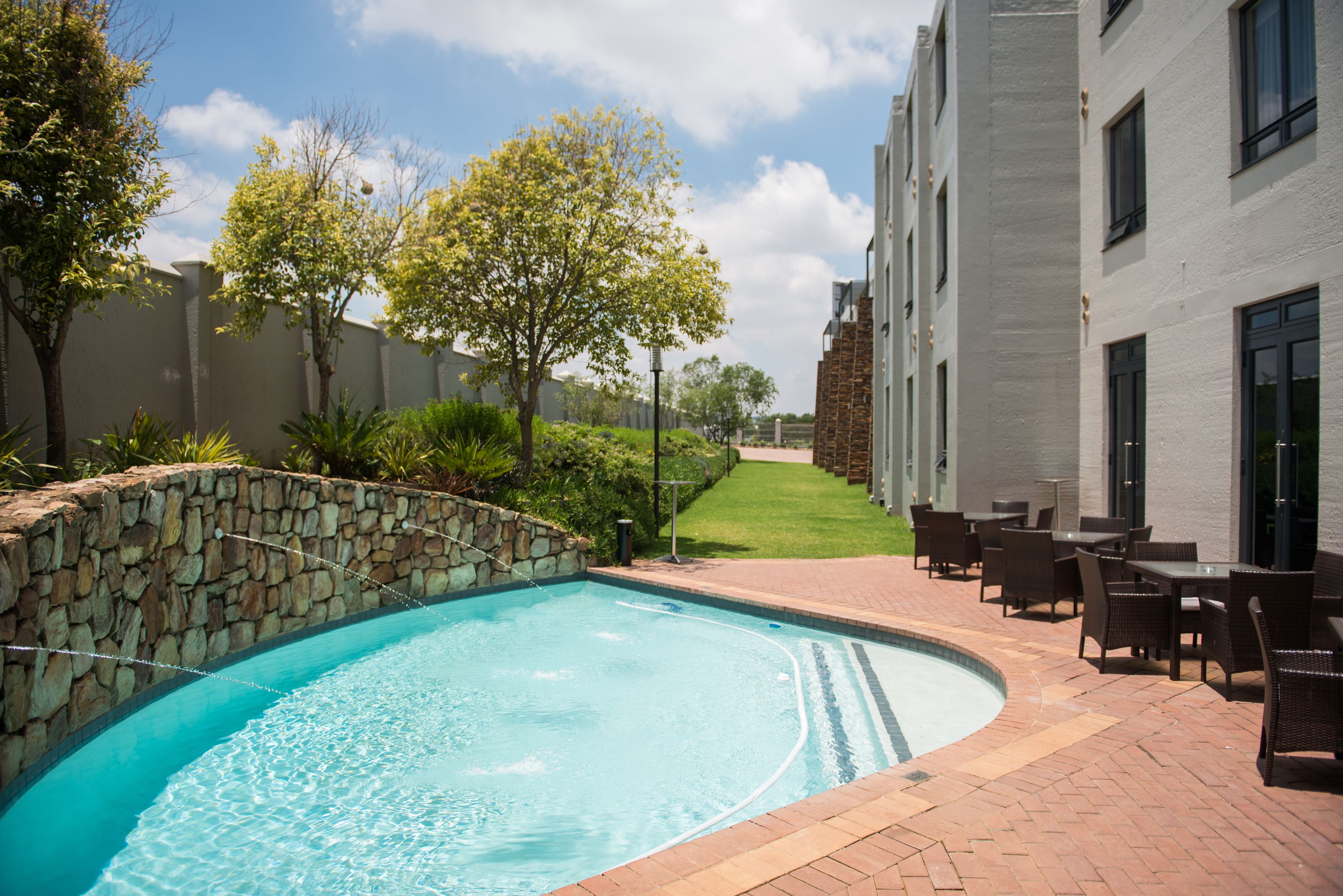 A swimming pool with a stone wall, surrounded by outdoor seating and a building with multiple windows.
