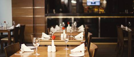 A restaurant with wooden tables set for dining, a bar area in the background, and a television screen.