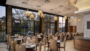 A spacious restaurant with multiple tables and chairs, large windows, and hanging pendant lights. - Premier Hotel Midrand (Midrand)