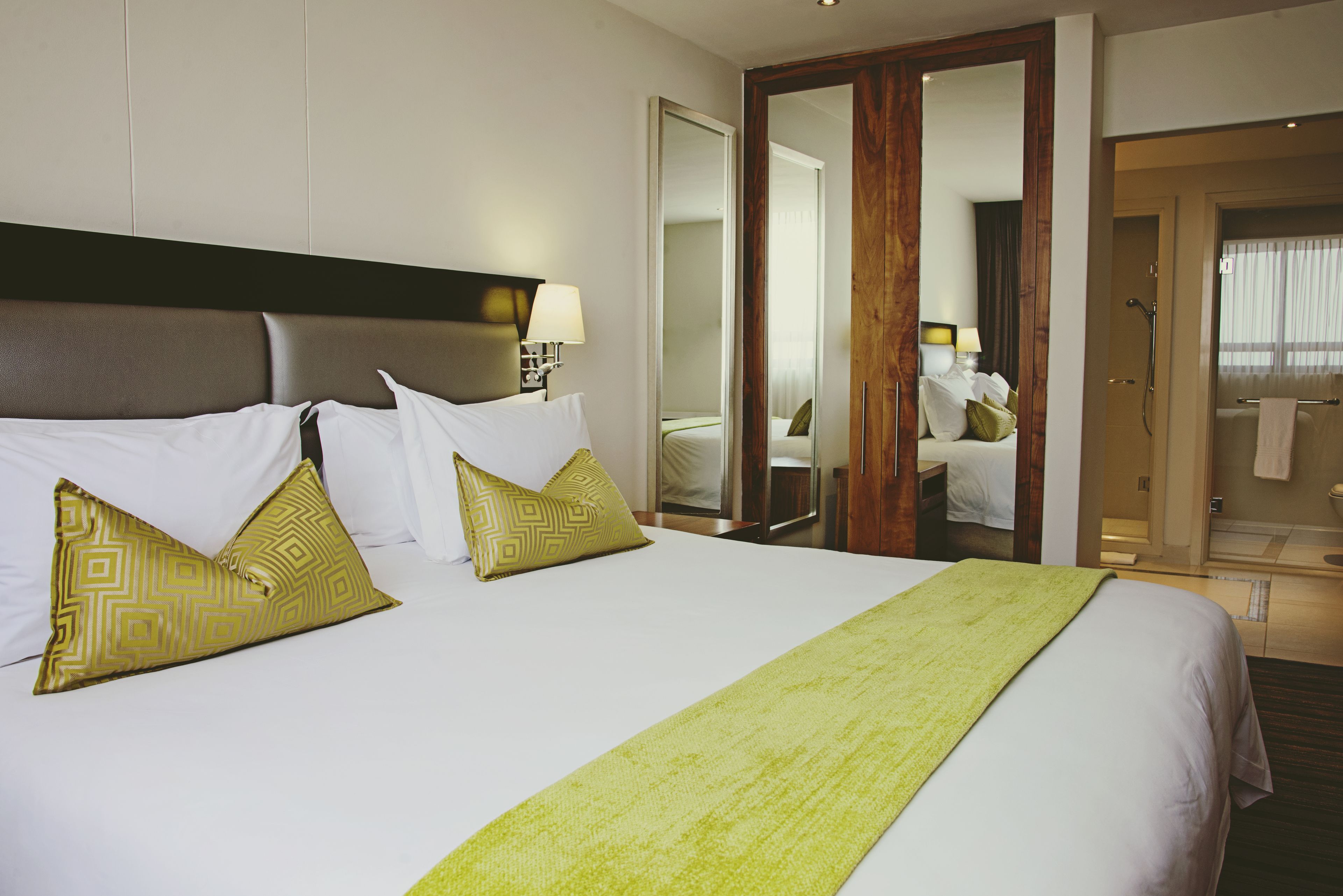 A hotel room with a large bed, two gold-patterned pillows, a green throw blanket, a wooden headboard, a mirror, and a bathroom visible through an open door.