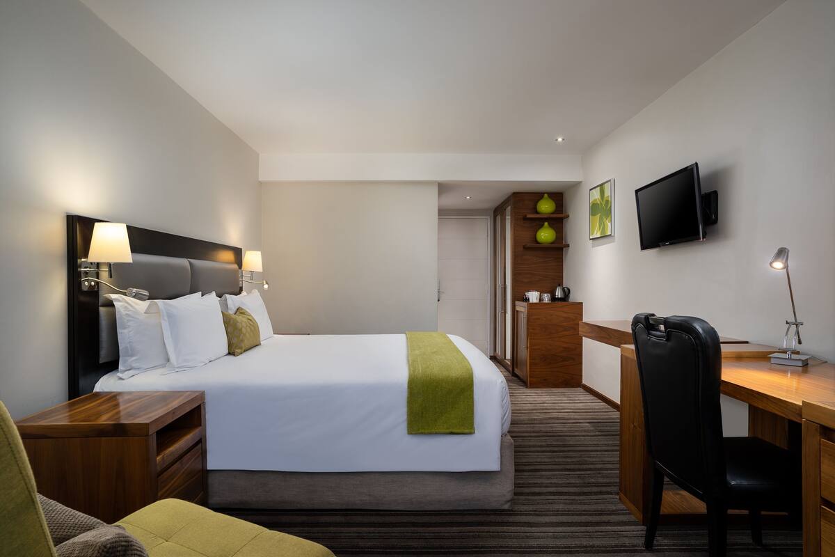 A hotel room with a bed, bedside tables, a desk with a chair, a TV, and a small kitchenette.