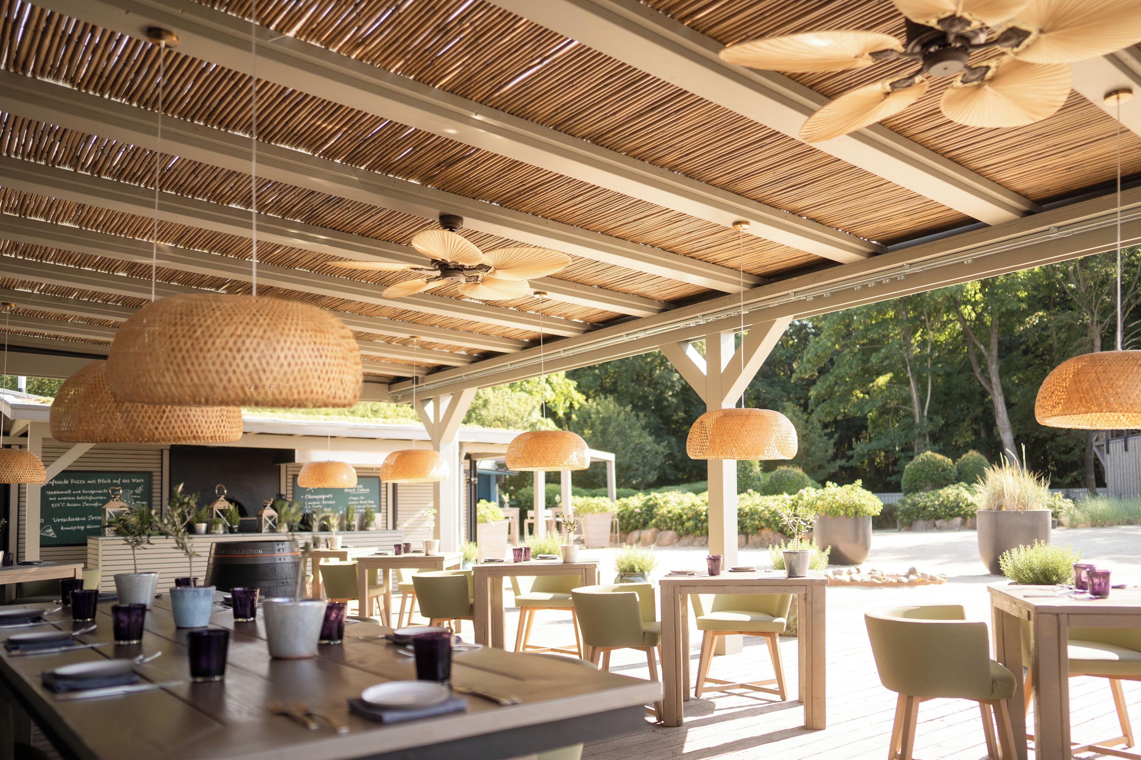 An outdoor dining area with wooden tables and chairs, hanging rattan lamps, and a view of greenery.