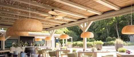 An outdoor dining area with wooden tables and chairs, hanging rattan lamps, and a view of greenery.