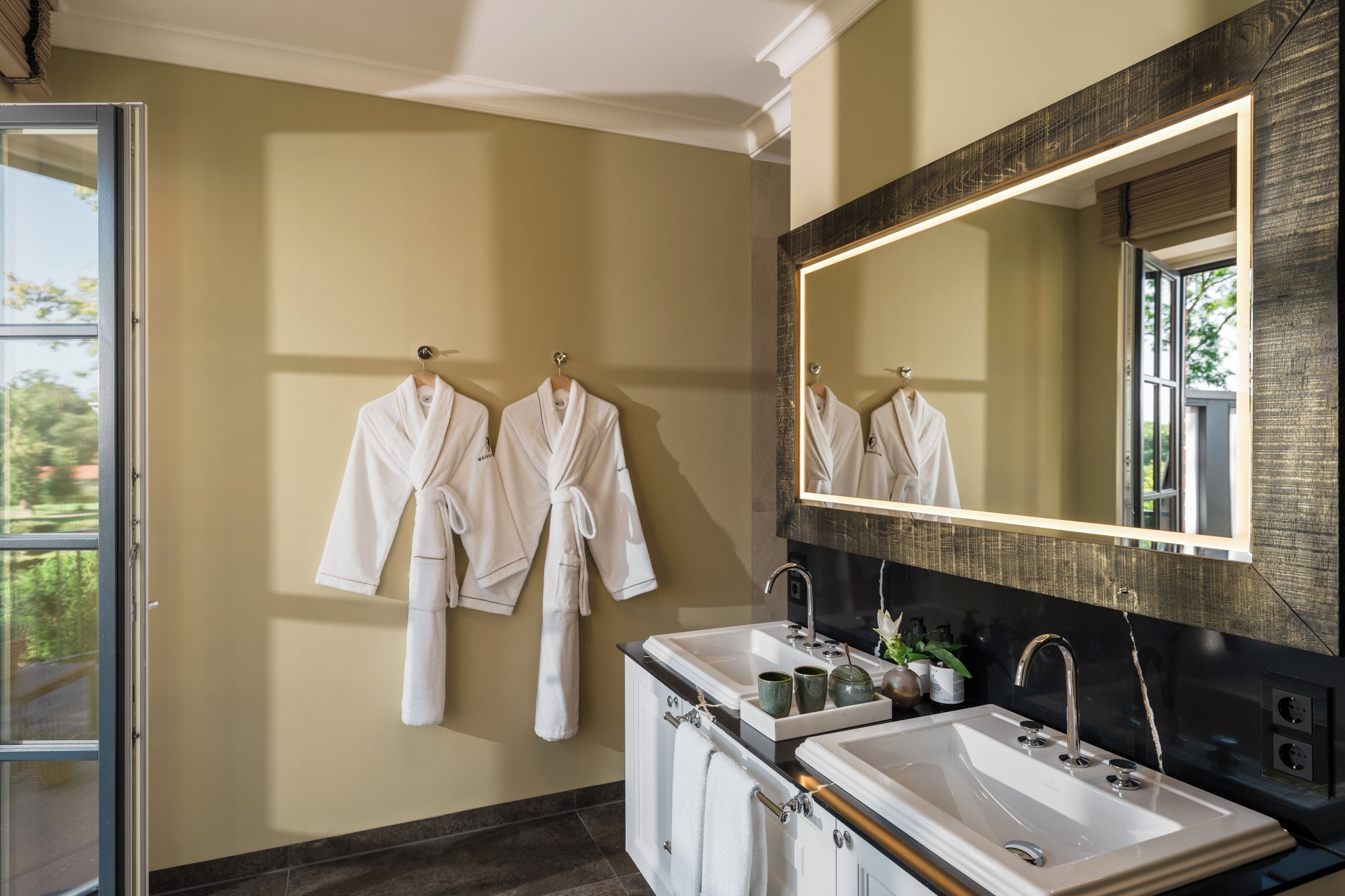 A bathroom with two sinks, a large mirror, and three white robes hanging on the wall.