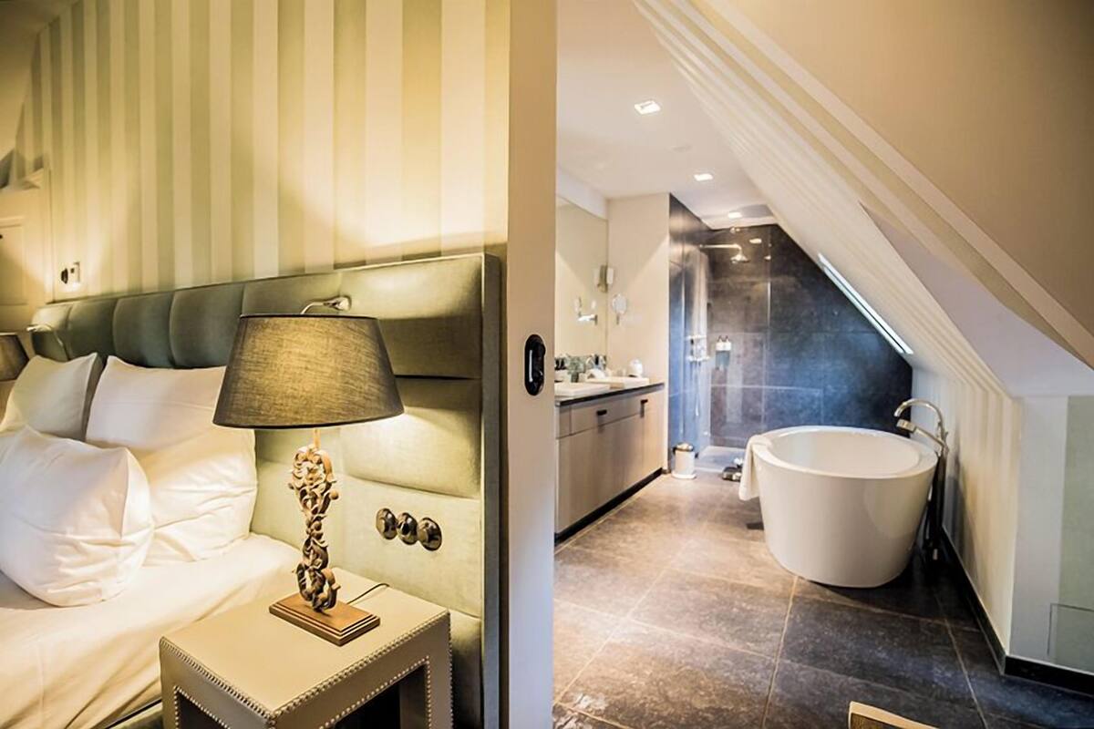 Une chambre à coucher avec un lit, une lampe de chevet et une table de nuit. La pièce est décorée de papier peint à rayures et le lit est doté d’une tête de lit. L’image montre également une salle de bain avec une baignoire, un lavabo et un miroir.