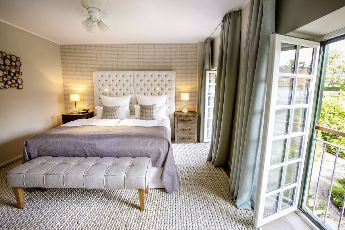 Une chambre avec un grand lit, deux tables de chevet, un banc et une vue sur l’extérieur grâce à une grande fenêtre.