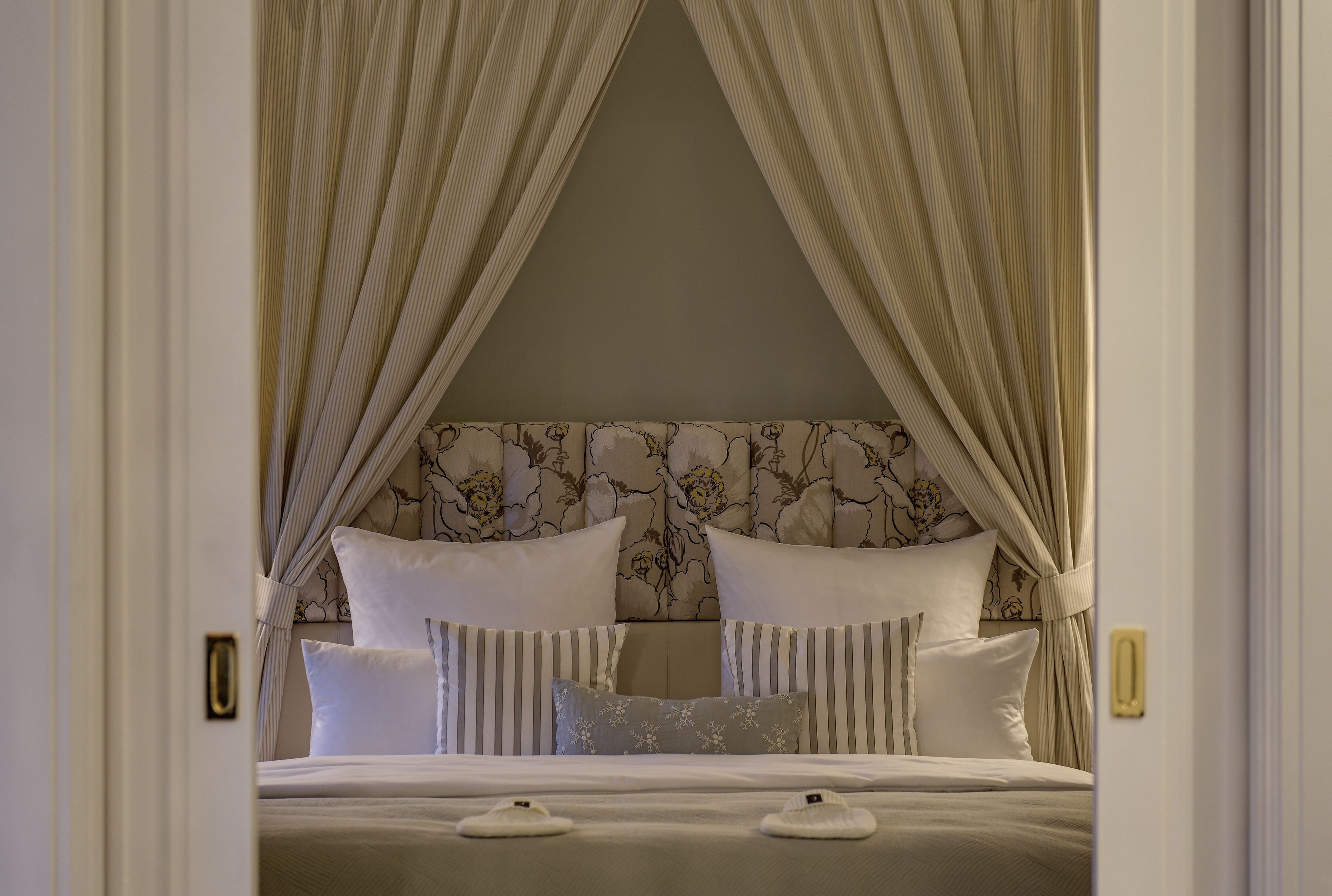 A neatly made bed with white bedding and striped pillows, a floral headboard, and sheer curtains.