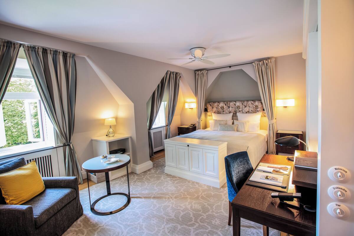Une chambre d’hôtel avec un lit, un bureau, une chaise, une petite table et une fenêtre avec des rideaux.