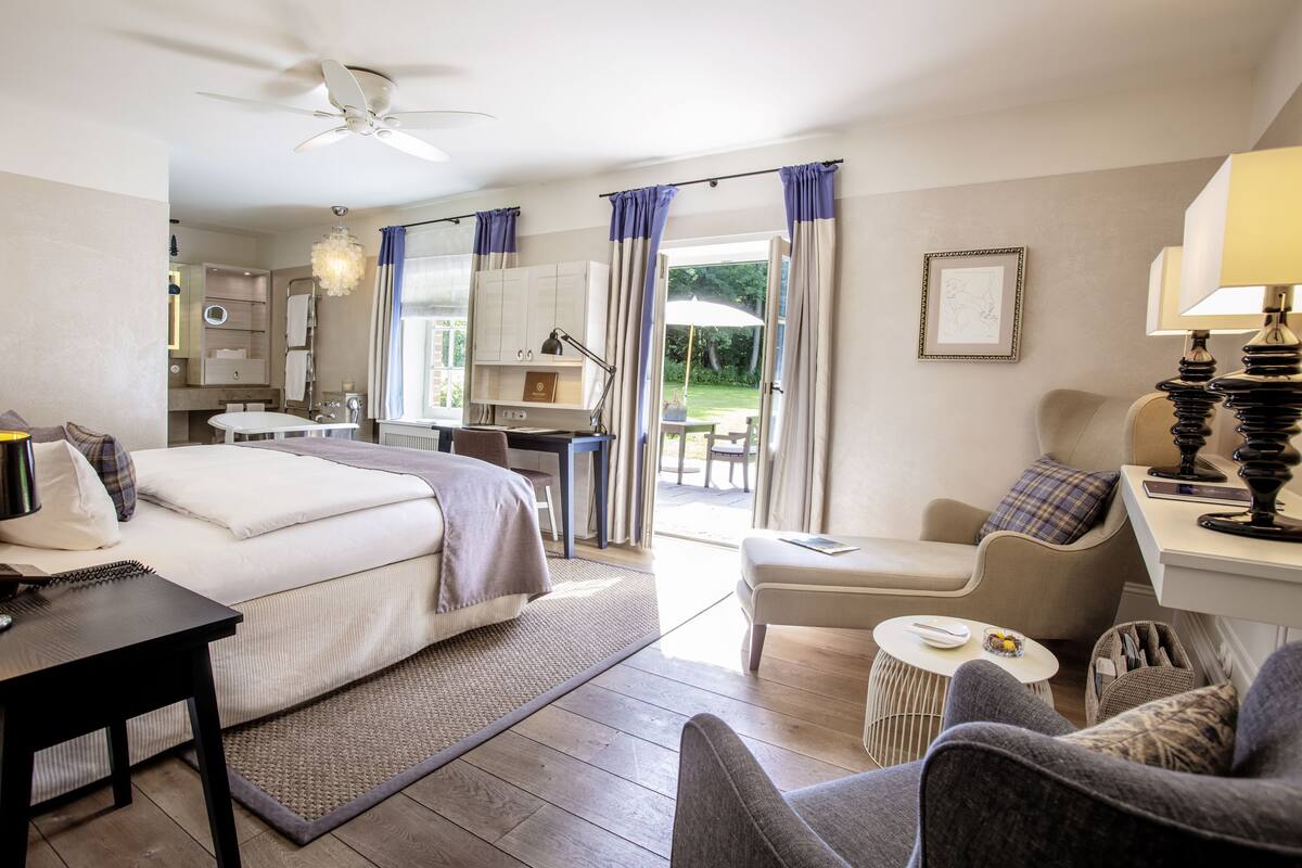 Une chambre bien éclairée, avec un lit, une table de chevet et un bureau avec une lampe. On peut voir un jardin par la fenêtre.