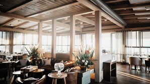 Een ruime eetruimte met houten balken, grote ramen en diverse zithoekjes, waaronder tafels en stoelen.