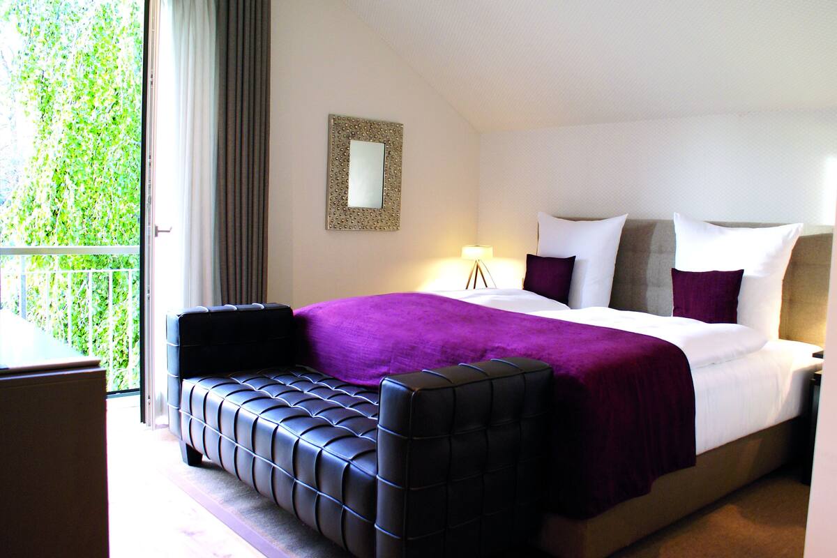 Une chambre d’hôtel avec un lit, un canapé noir, un balcon donnant sur un paysage verdoyant et un miroir fixé au mur.