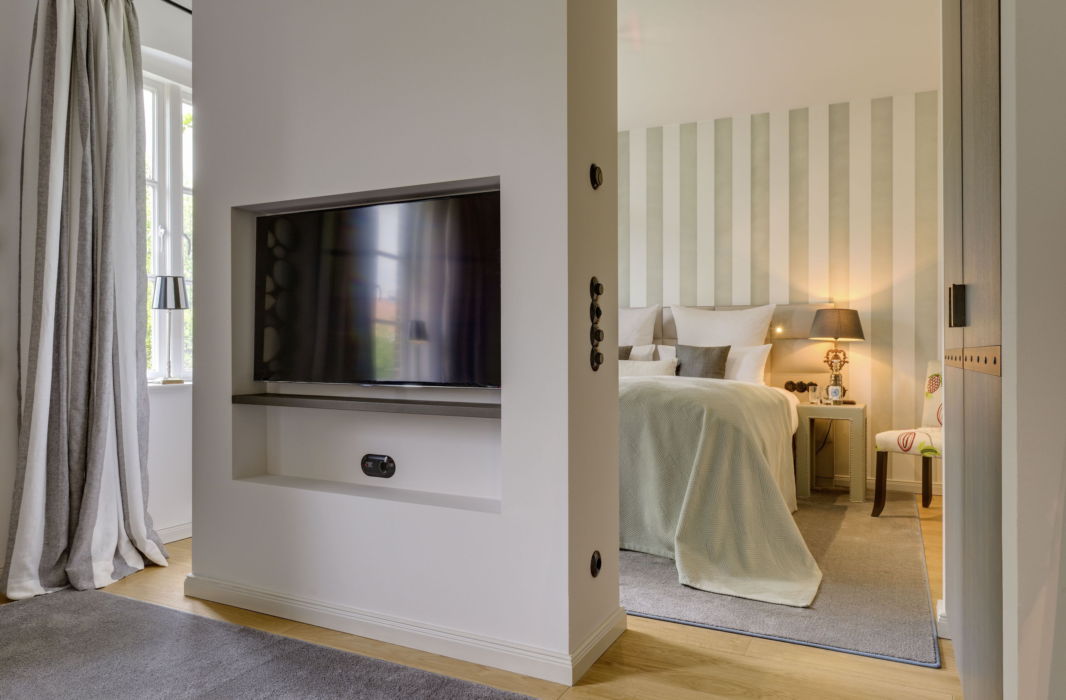 Ein modernes Schlafzimmer mit einem an der Wand montierten Flachbildfernseher, einem Bett mit grüner Decke, einem Nachttisch mit Lampe und einem Sessel.