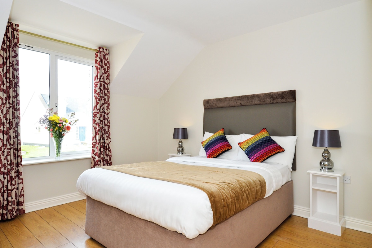 Una cama bien tendida con una colcha color crema y dos almohadas coloridas, una ventana con cortinas estampadas y una mesita de noche con una lámpara.