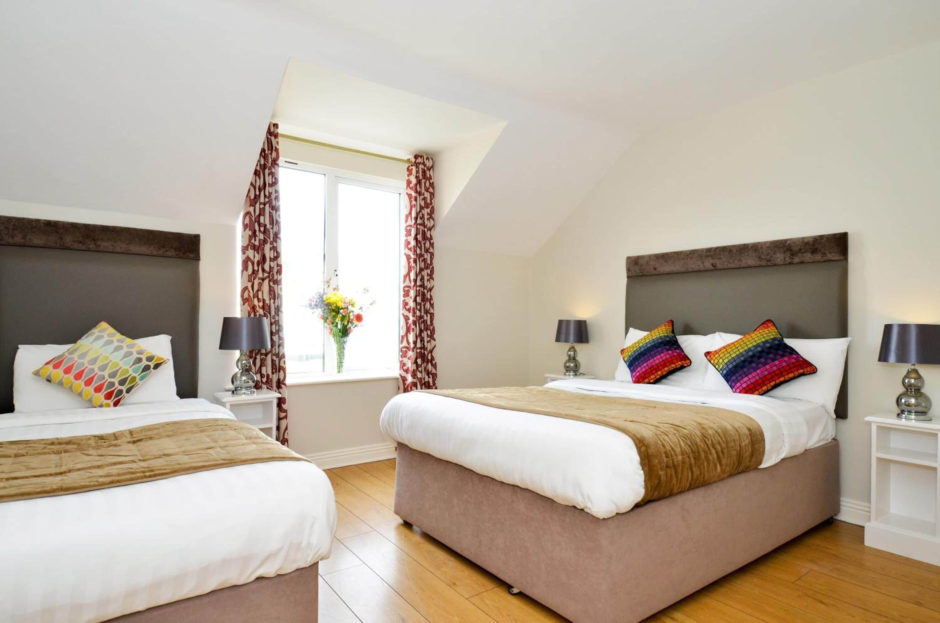 A hotel room with two beds, a window with curtains, a vase with flowers, and a lamp on each nightstand.