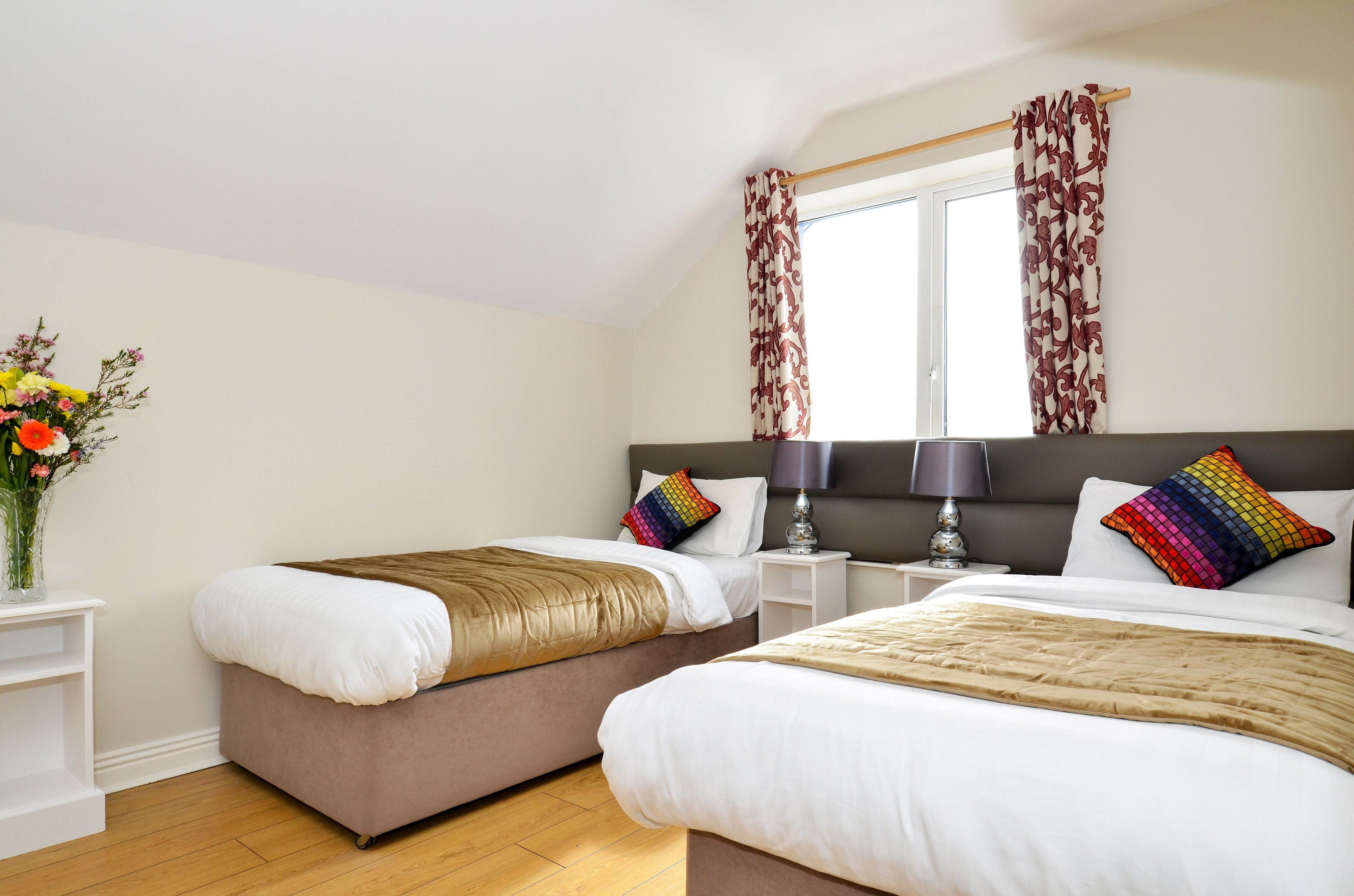 A bedroom with two beds, a window with patterned curtains, a bedside table with a lamp, and a vase with flowers.